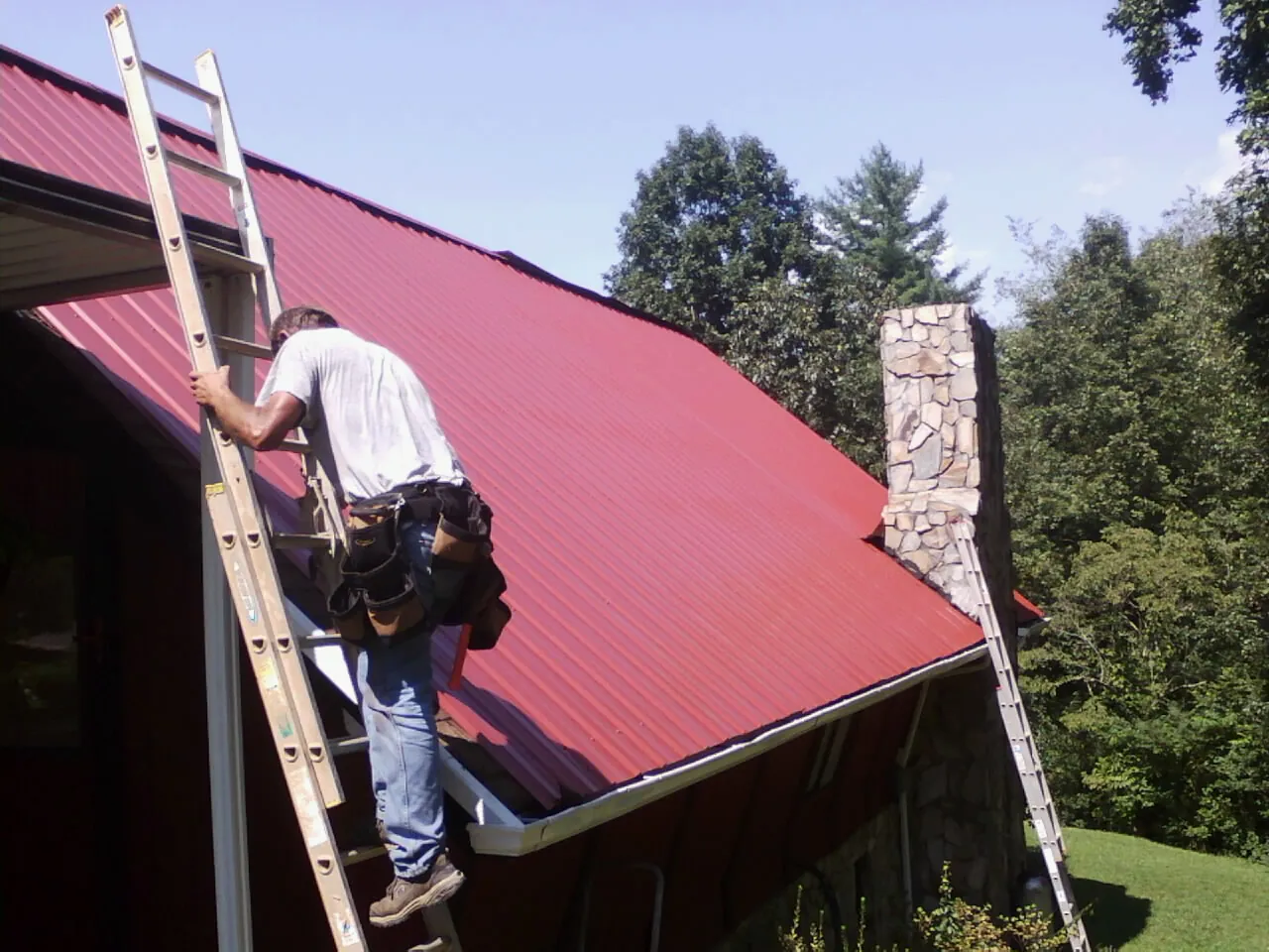 Roofer on a ladder beside a red metal roof and stone chimney — Soffit & Fascia Repair by Roofing Brothers in Mackville
